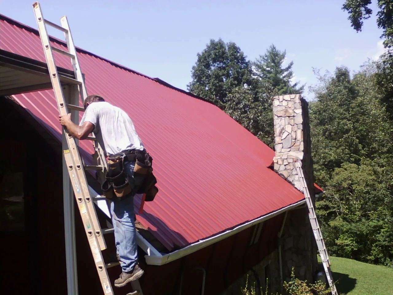 Roofer on a ladder beside a red metal roof and stone chimney — Soffit & Fascia Repair by Roofing Brothers in Lake Andes